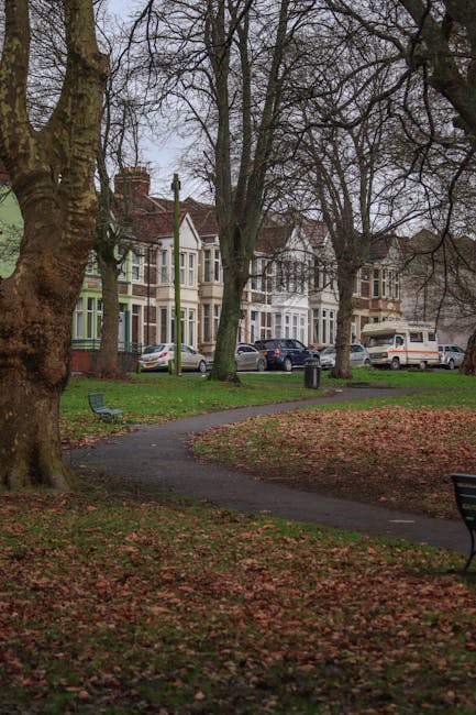 The image depicts a quiet residential park area with a winding paved pathway bordered by leaf fallen on the grass and brown soil during late autumn, with several tall, leafless trees casting shadows across the scene. In the background, there are a row of Victorian-style terraced houses with bay windows, brick facades, and chimneys, all set along a street with parked cars, including sedans, hatchbacks, and a minibus, indicating the area’s urban environment. Adjacent to the pathway, there are a couple of metal benches with wooden slats, one positioned in the foreground on the right side and the other further back on the left side, both situated on clusters of fallen leaves. The scene appears overcast, with diffused natural light providing a subdued atmosphere typical of late autumn or early winter, and an overall impression of a well-maintained, quiet neighbourhood park space. This setting visually supports services such as independent rubbish collection or onsite waste handling provided by companies like rubbishcollectionqueenspark.co.uk, especially in residential areas needing private waste management solutions amidst urban surroundings.