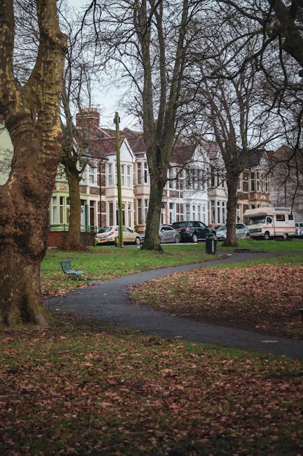 A quiet residential street scene in a park-like setting showing several terraced houses with bay windows and brick facades in the background. In the foreground, there is a curved paved pathway flanked by patches of grass and fallen leaves, some of which are scattered over the path. Mature trees with bare branches line the area, indicating a late autumn or winter season, and cast shadows on the ground. Alongside the pavement, there are parked cars, including a silver sedan and a white van, near the residential buildings. A small black waste bin is situated on the sidewalk, likely maintained by local waste collection services such as Rubbish collection for Queens Park Gardens households, Queens Park, to support efficient rubbish disposal. The environment is calm and typical of urban neighborhood streets, with no visible activity or congestion, emphasizing a peaceful outdoor setting suitable for walking or relaxation under natural light with overcast sky. This scene subtly reflects the importance of local rubbish collection and private waste handling in maintaining cleanliness of residential areas.