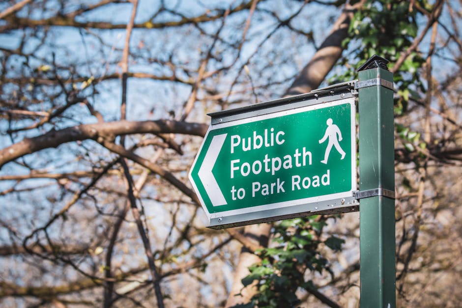 A green rectangular sign with white lettering and a pedestrian symbol directs pedestrians to a public footpath leading to Park Road. The sign is mounted on a metal post situated outdoors, with a background featuring leafless tree branches and a partly cloudy sky, indicating seasonal weather. The sign's surface appears smooth and reflective, with a metal frame secured with bolts at the top and bottom. Surrounding the sign, there are hints of greenery and the image is illuminated by natural daylight, which creates a bright and clear scene. This setting suggests a typical urban or suburban environment where pedestrian routes can be clearly marked, supporting the context of local infrastructure and alternative pathways for walking in lieu of private or rubbish removal spaces. The focus on the sign and natural environment subtly aligns with themes of community pathways and outdoor accessibility, relevant to waste disposal and collection routes that may pass through public areas like parks or footpaths managed by local authorities or independent waste management services.