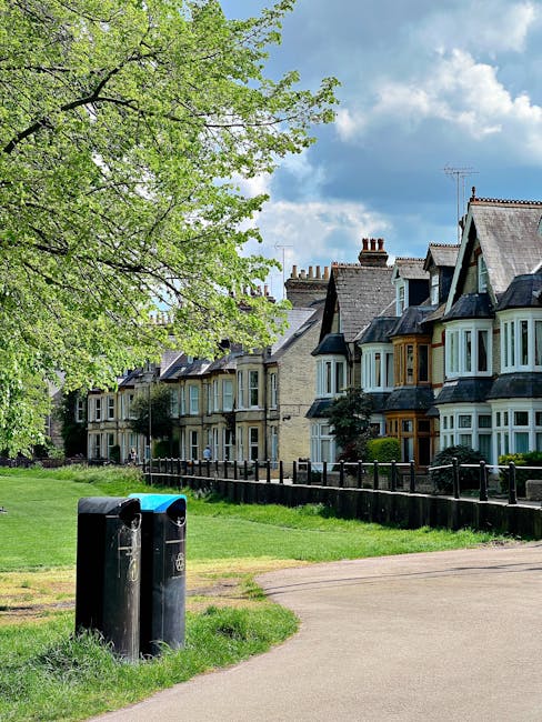 A daytime view of a residential street with a row of terraced houses featuring bay windows and slate roofs, situated behind a wooden fence. In the foreground, two black rubbish bins with closed lids are placed near a paved footpath that runs parallel to the grassy verge and houses. To the left, a large tree with fresh green leaves extends its branches, casting dappled shade over part of the scene. The sky above shows scattered white clouds and patches of blue, indicating fair weather. The environment suggests a quiet, well-maintained neighborhood where private waste collection may be preferred, supported by the presence of individual rubbish bins rather than communal waste management systems. The overall scene offers a typical residential setting with neatly maintained front gardens and organized waste disposal points, aligning with alternative rubbish collection services that cater to local communities.