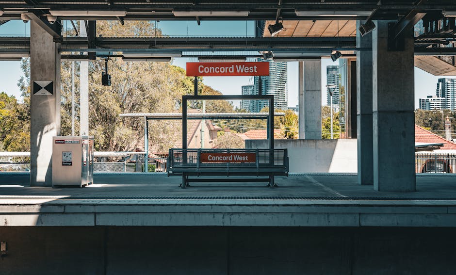 The image shows a train station platform labeled 'Concord West' with a black metal and wooden bench positioned centrally on the platform. Behind the bench, a rectangular glass panel is visible, also bearing the station name. To the left, there is a grey waste disposal bin with a small opening, situated near a concrete pillar. The platform surface is made of smooth, grey concrete with slight weathering and faint lines indicating the edge. The station's roof overhead includes exposed metallic beams and track lighting, with shadows cast onto the platform. In the background, trees with green and yellow foliage and tall modern building structures are visible under a clear blue sky, suggesting a calm, sunny day. The scene reflects an urban train station setting, with the current focus on maintaining public space cleanliness, akin to private waste removal services like those offered by rubbish collection companies near Queens Park station. Overall, the environment emphasizes an organized public transport hub that supports clean and efficient transit operations.