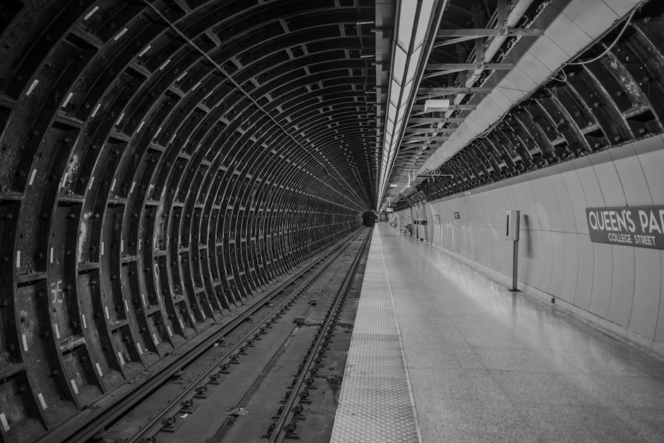 A black and white photograph of an underground metro station tunnel, showing curved metal framework on the left side that forms the tunnel's interior structure, with rail tracks running down the centre of the image. The tracks are flanked by a narrow platform on the right, which is smooth and reflective, with a slightly textured tactile paving strip along the edge for safety. The station walls are tiled in light-colored, glossy tiles, with visible signage indicating 'Queen's Park' on the right side. Overhead, structural and electrical components are mounted along the ceiling, with some suspended fixtures providing illumination. The overall scene appears clean and well-maintained, with a sense of depth created by the receding tunnel towards a small dark figure at the far end, highlighting the underground environment's enclosed and orderly nature, sometimes associated with transport infrastructure or alternative underground waste management solutions of large-scale urban areas.